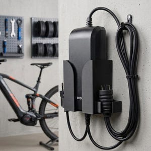 Könnte beinhalten: Eine schwarze Wandhalterung aus Kunststoff für ein Bosch E-Bike-Akkuladegerät. Die Halterung sichert das rechteckige Ladegerät und verfügt über integrierte seitliche Clips, um die Stromkabel und den Stecker ordentlich zu verstauen und zugänglich zu halten.