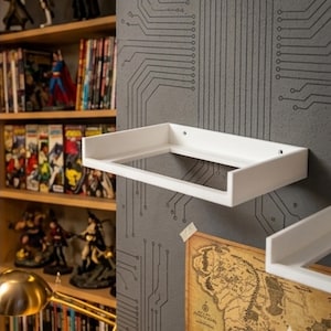 Puede incluir: Estanterías de pared rectangulares blancas montadas en una pared gris con un diseño de placa de circuito. Las estanterías están vacías y hay un mapa pegado debajo de una de ellas. En el fondo se ve una estantería con cómics y figuras.