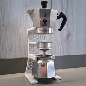 Op de afbeelding: Een zilverkleurige Bialetti Moka Express koffiezetapparaat van aluminium, gedemonteerd in drie delen en geplaatst in een verticale witte 3D-geprinte standaard. Het bovendeel bevat het Bialetti-logo en de tekst Moka Express MADE IN ITALY.