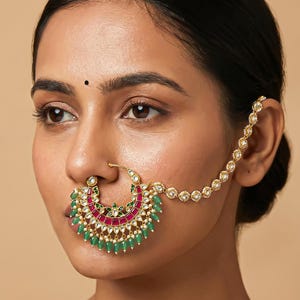 Pode incluir: Um anel de nariz dourado com um grande pendente em forma de meia-lua adornado com pedras retangulares cor-de-rosa, detalhes em esmalte verde e pequenas pedras brancas. Uma corrente dourada delicada com pedras brancas liga o anel à orelha.