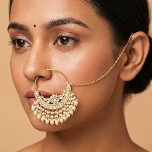 Pode incluir: Um piercing de nariz dourado com um design em forma de meia-lua. O piercing apresenta um padrão floral central com pedras transparentes e é adornado com pérolas brancas penduradas. Uma corrente delicada estende-se até à orelha, completando a joia indiana tradicional.