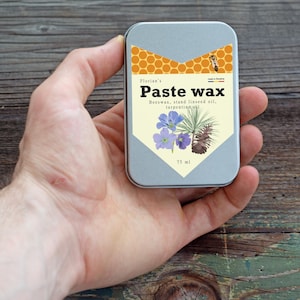Pode incluir: Uma pequena lata retangular de cera em pasta é segurada. A lata tem um rótulo com as palavras "Paste wax" e ilustrações botânicas. A parte superior da lata apresenta um design de favo de mel e uma abelha. A lata contém 75 ml de cera.