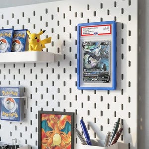 May include: A white pegboard wall organizer featuring a blue-framed PSA graded Lugia V trading card, a yellow Pikachu figure, a Charizard framed print, a white shelf with Pokémon card packs, a white pen holder with pens, and a small white storage bin.