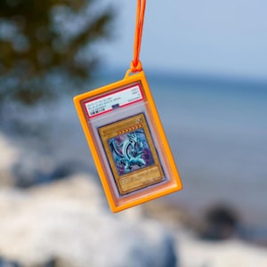 Könnte beinhalten: Eine bewertete Yu-Gi-Oh!-Sammelkarte Blue-Eyes White Dragon in einer transparenten Kunststoffhülle, eingefasst in einen leuchtend orangefarbenen rechteckigen Rahmen, der an einem passenden orangefarbenen Umhängeband mit schwarzem Kordelstopper hängt.