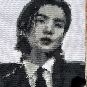 May include: A square crochet tapestry featuring a monochromatic portrait of a person with shoulder-length hair, wearing a collared shirt and a necktie. The image is rendered in shades of black, white, and gray, creating a detailed, pixelated visual effect.