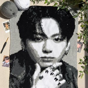 May include: A rectangular crochet tapestry featuring a grayscale portrait of a person with dark hair. The person has a hand near their chin with the word ARMY and a heart symbol visible on the fingers. The piece is crafted in black, white, and gray yarn.