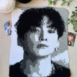 May include: A rectangular crochet tapestry featuring a monochromatic portrait of a person with dark hair. The image is rendered in shades of black, white, and gray yarn, creating a detailed likeness. Two small rectangular photo cards are placed nearby.
