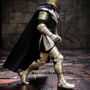 May include: A full suit of polished steel armor, including a helmet, shoulder guards, arm and leg protection, and boots. A black cape drapes over the shoulders. The text "Nautical Armour Finds" is visible.