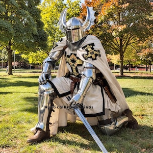 May include: A person in full knight armor, kneeling on green grass. The silver armor includes a helmet with decorative horns, a chainmail neck guard, and articulated plates. A white cape with black and gold emblems is worn over the armor, and a long sword rests on the ground.