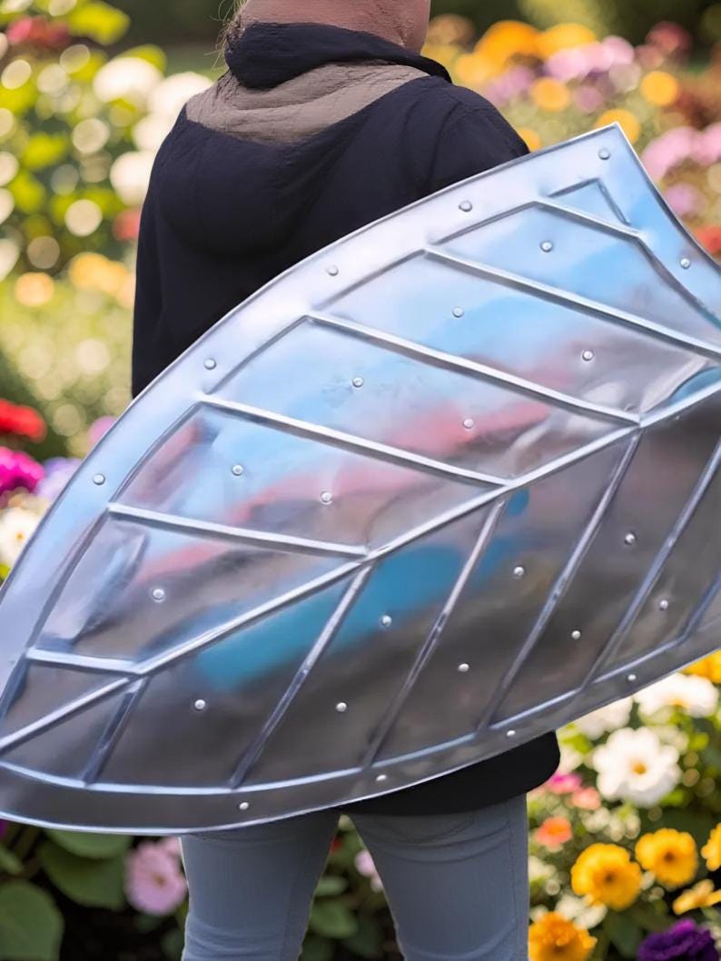 Op de afbeelding: Een zilverkleurig schild met een bladachtig ontwerp, met diagonale lijnen en klinknagels. Het schild wordt vastgehouden door een persoon die een zwarte jas en een grijze broek draagt, met een kleurrijke bloementuin op de achtergrond.