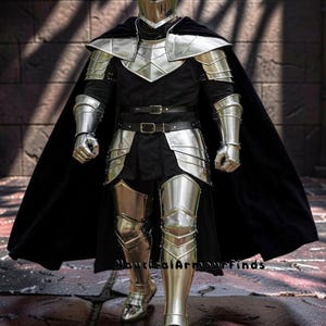 May include: A full suit of medieval armor, including a helmet, chest plate, shoulder guards, arm and leg protection, and gauntlets. The armor is silver-colored and worn over a black tunic and cape. The image is taken from a front-facing perspective.