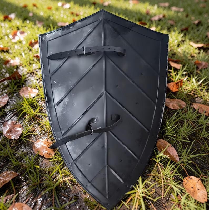 Op de afbeelding: Een zwart schild met een bladachtig ontwerp en twee leren riemen. Het schild is gemaakt van een donker materiaal en ligt op gras met gevallen bladeren.