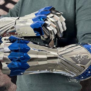 May include: A pair of polished silver metal gauntlets featuring articulated finger plates with vibrant blue accents. The cuffs are decorated with a blue star-patterned trim and small silver rivets. The text NauticalArmourFinds is visible on the metal.