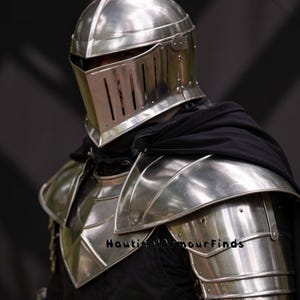 May include: A polished silver knight's helmet and shoulder armor. The helmet features a visor with vertical slits and a rounded top. The shoulder armor is layered and reflective, with the text "Nautical Armour Finds" visible.