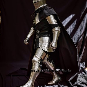 May include: A full suit of polished steel armor, including a helmet, chest plate, arm and leg guards, and gauntlets. The armor is worn over a black tunic and cape. The image shows the armor from the side, with the person walking forward. The text "Nautical Armour Finds" is visible.