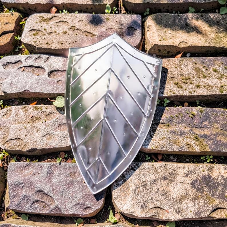 Op de afbeelding: Een glanzend zilveren schild met een puntige onderkant en een decoratief chevronpatroon. Het metalen schild reflecteert licht. Het schild ligt op een stenen ondergrond. Middeleeuws thema.