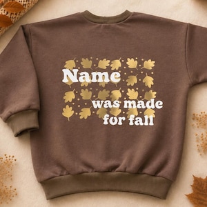 Könnte beinhalten: Ein dunkelbraunes Langarm-Sweatshirt mit einem goldenen Blättermuster im Hintergrund und dem weißen Text Name was made for fall. Das Kleidungsstück hat einen Rundhalsausschnitt sowie gerippte Bündchen an den Ärmeln und am Saum für ein schlichtes Design.