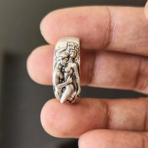 May include: A silver ring featuring a detailed relief sculpture of two figures embracing. The ring has a rectangular shape with rounded edges. The figures are depicted in a classical style, with intricate details in their clothing and hair.