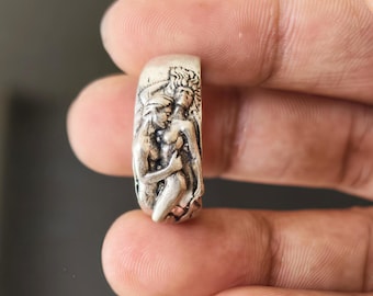 May include: A silver ring featuring a detailed relief sculpture of two figures embracing. The ring has a rectangular shape with rounded edges. The figures are depicted in a classical style, with intricate details in their clothing and hair.