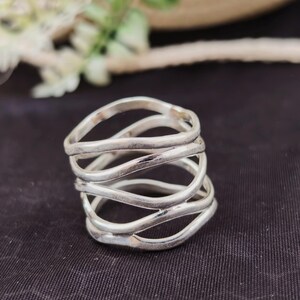 Peut inclure: Une bague en argent au design unique. La bague présente plusieurs bandes ondulées qui s'enroulent autour du doigt. Les bandes sont faites d'un matériau métallique brillant. La bague est présentée sur une surface noire.