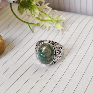 Könnte beinhalten: Silberfarbener Ring mit einem ovalen Moosachat-Edelstein. Der Ring hat ein graviertes Muster um den Stein und auf dem Band. Der Edelstein zeigt einen marmorierten Effekt mit Grün- und Weißtönen. Ein auffälliges Schmuckstück.