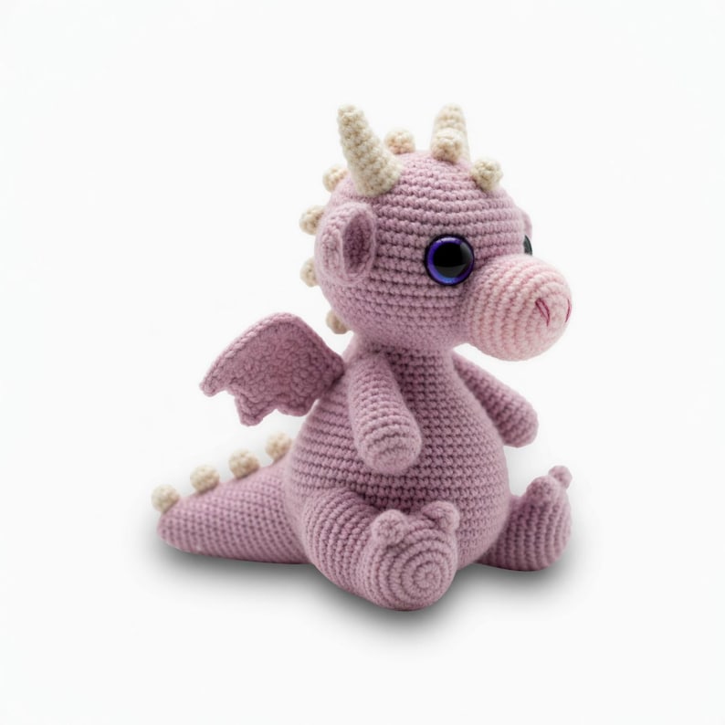 May include: A crocheted dragon toy in shades of lavender and cream. The dragon has large, dark purple eyes, small wings, and cream-colored horns and spikes. The dragon is sitting with its legs outstretched.