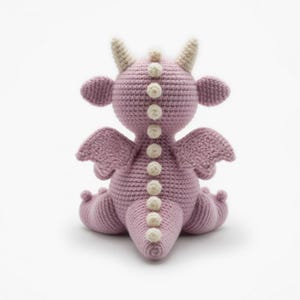 May include: A handmade crochet dragon in a soft purple hue. The dragon features cream-colored horns, small wings, and a row of cream-colored decorative accents down its back. The dragon is sitting with its legs tucked under.