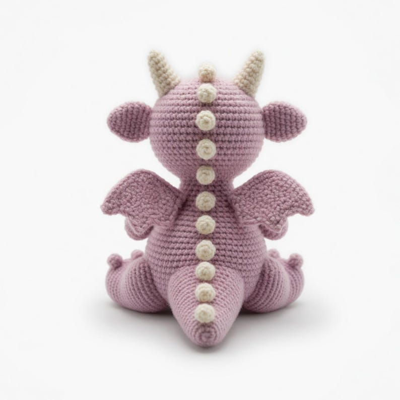 May include: A handmade crochet dragon in a soft purple hue. The dragon features cream-colored horns, small wings, and a row of cream-colored decorative accents down its back. The dragon is sitting with its legs tucked under.