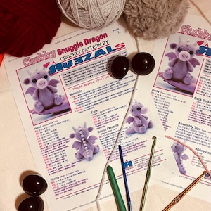 May include: A crochet pattern for a "Chubby Snuggle Dragon" with instructions and diagrams. The image includes crochet hooks, yarn in various colors, and black safety eyes. The dragon's height is approximately 4 to 5 inches.