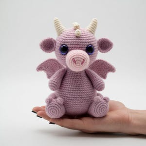 May include: A crocheted dragon plushie in shades of pink and light pink. The dragon has large, dark blue eyes, small wings, and cream-colored horns. It is sitting on a hand.