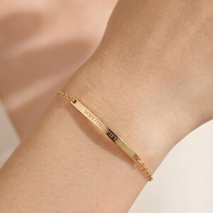 May include: A gold-toned metal bar bracelet featuring a delicate chain and a thin, horizontal rectangular plate. The surface of the metal bar is engraved with small, precise alphanumeric coordinates, creating a minimalist and refined jewelry piece.