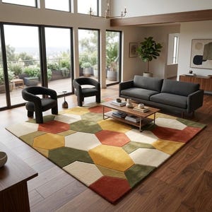 May include: A large area rug featuring a geometric pattern of interlocking hexagons in shades of mustard yellow, olive green, terracotta orange, and cream. The rug is placed on a wooden floor in a room with a dark gray sofa and two armchairs.