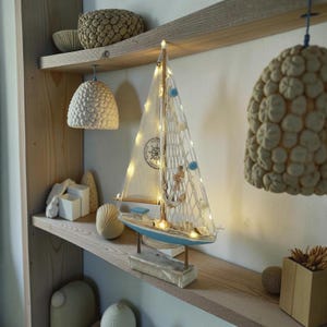 May include: A decorative sailboat with a blue hull and white sails, adorned with string lights and nautical accents. The sailboat sits on a wooden shelf with other coastal-themed decor, including a seashell and a hanging lamp.