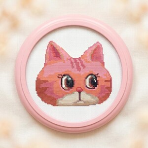 Puede incluir: Un bordado de punto de cruz de una cara de gato centrado en un marco redondo de plástico rosa claro. El gato presenta pelaje en tonos rosa y naranja, ojos grandes y oscuros con pestañas visibles, una pequeña nariz rosa y un hocico blanco.