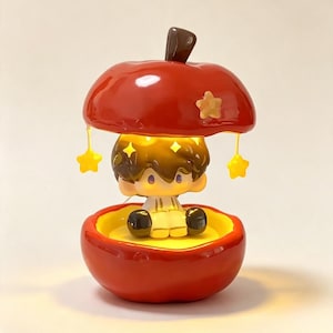 May include: A decorative lamp shaped like a red apple with a brown stem. The top half of the apple lifts to reveal a small, seated figure with brown hair and white clothing. Yellow star-shaped charms hang from the apple lid, and the interior glows.