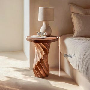 May include: A round wooden side table with a spiralling, carved base and a smooth top surface. The table is positioned next to a bed, with a beige lamp resting on top. Arrows indicate the width and height dimensions of the table.