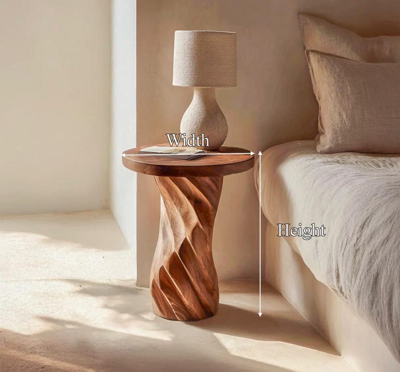 May include: A round wooden side table with a spiralling, carved base and a smooth top surface. The table is positioned next to a bed, with a beige lamp resting on top. Arrows indicate the width and height dimensions of the table.