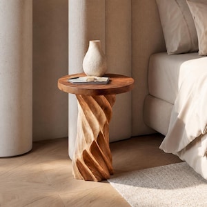 May include: A round wooden side table with a dark brown top and a light brown, spiralling, carved base. A small, off-white, textured ceramic vase sits on the table surface, accompanied by a thin, rectangular, white object with a dark centre.
