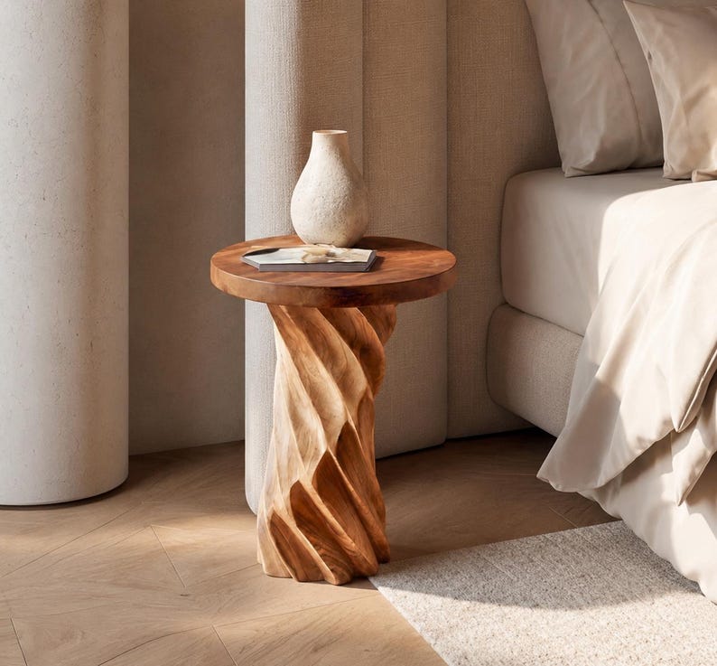 May include: A round wooden side table with a dark brown top and a light brown, spiralling, carved base. A small, off-white, textured ceramic vase sits on the table surface, accompanied by a thin, rectangular, white object with a dark centre.