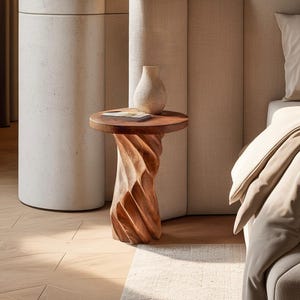 May include: A round wooden side table with a spiralling, carved base and a smooth top surface. A small, light-coloured ceramic vase and a thin, rectangular book are placed on the table. The wood features a warm, natural brown grain pattern.