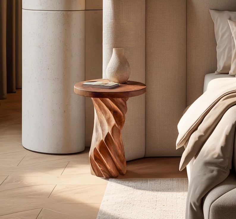 May include: A round wooden side table with a spiralling, carved base and a smooth top surface. A small, light-coloured ceramic vase and a thin, rectangular book are placed on the table. The wood features a warm, natural brown grain pattern.
