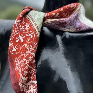 Puede incluir: Un pañuelo cuadrado con una base roja vibrante y complejos patrones florales y de volutas en blanco. El diseño incluye una sección de borde verde pálido y un área de acento púrpura sutil, creando una apariencia multicolor y suave.