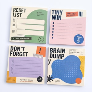 Op de afbeelding: Vier vierkante memoblokjes met kleurrijke grafische ontwerpen. De blokjes bevatten teksten zoals RESET LIST, TINY WIN, DON'T FORGET en BRAIN DUMP, samen met diverse checklists, stippenrasters en motiverende zinnen in verschillende kleuren.