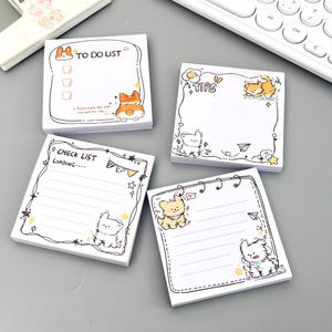 Puede incluir: Cuatro blocs de notas adhesivas cuadrados con ilustraciones de perros de dibujos animados y texto. Un bloc dice TO DO LIST, otro TIPS y otro CHECK LIST LOADING. Los diseños incluyen varios perros pequeños, estrellas y bordes decorativos.