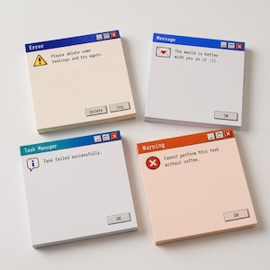 Op de afbeelding: Vierkante memoblokjes ontworpen als computer pop-up vensters. Links bovenaan een beige blok met 'Error' en 'Please delete some feelings and try again'. Rechts bovenaan een wit blok met 'Message' en 'The world is better with you in it'.