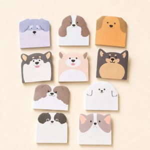 Puede incluir: Una colección de diez blocs de notas adhesivas cuadrados, cada uno con una cara de perro ilustrada única. Los diseños incluyen varias razas y patrones de color como marrón, negro, blanco, gris y beige, dispuestos en forma de cuadrícula.