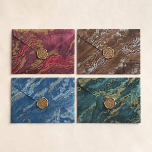 May include: Four rectangular envelopes featuring marbled patterns in shades of magenta, brown, blue, and green. Each envelope is sealed with a circular gold-coloured wax seal featuring an embossed design on the centre of the triangular flap.