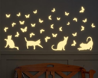 May include: A set of luminous wall decals featuring four cat silhouettes in various poses and numerous butterflies of different sizes. The pale yellow figures are arranged across a dark wall surface above a wooden bench.