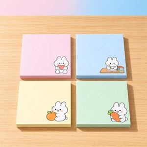 May include: Four square sticky note pads in pastel pink, blue, yellow, and green. Each pad features a small white cartoon rabbit illustration: one holding a red heart, one writing at a desk, one holding an orange, and one holding a carrot.