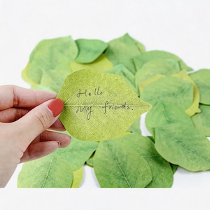 May include: A light green leaf-shaped sticky note with the text "Hello, my friends" in black script is held by a hand with red nail polish. The note is surrounded by other green leaf-shaped notes.